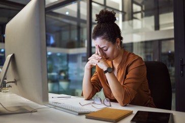 A young woman is at her desk in front of a large desktop computer; her head is down in exhaustion.