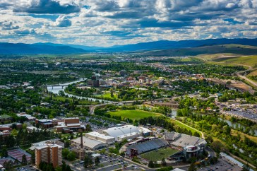 A skyline view of Missoula, Montana; buildings between rolling green hills give way to big mountains in the distance.