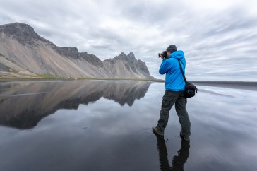 A man in a blue hoodie holds his camera up to a mountainscape; the sky is gray, and the ground seems to be wet sand, reflecting the mountains and the sky.