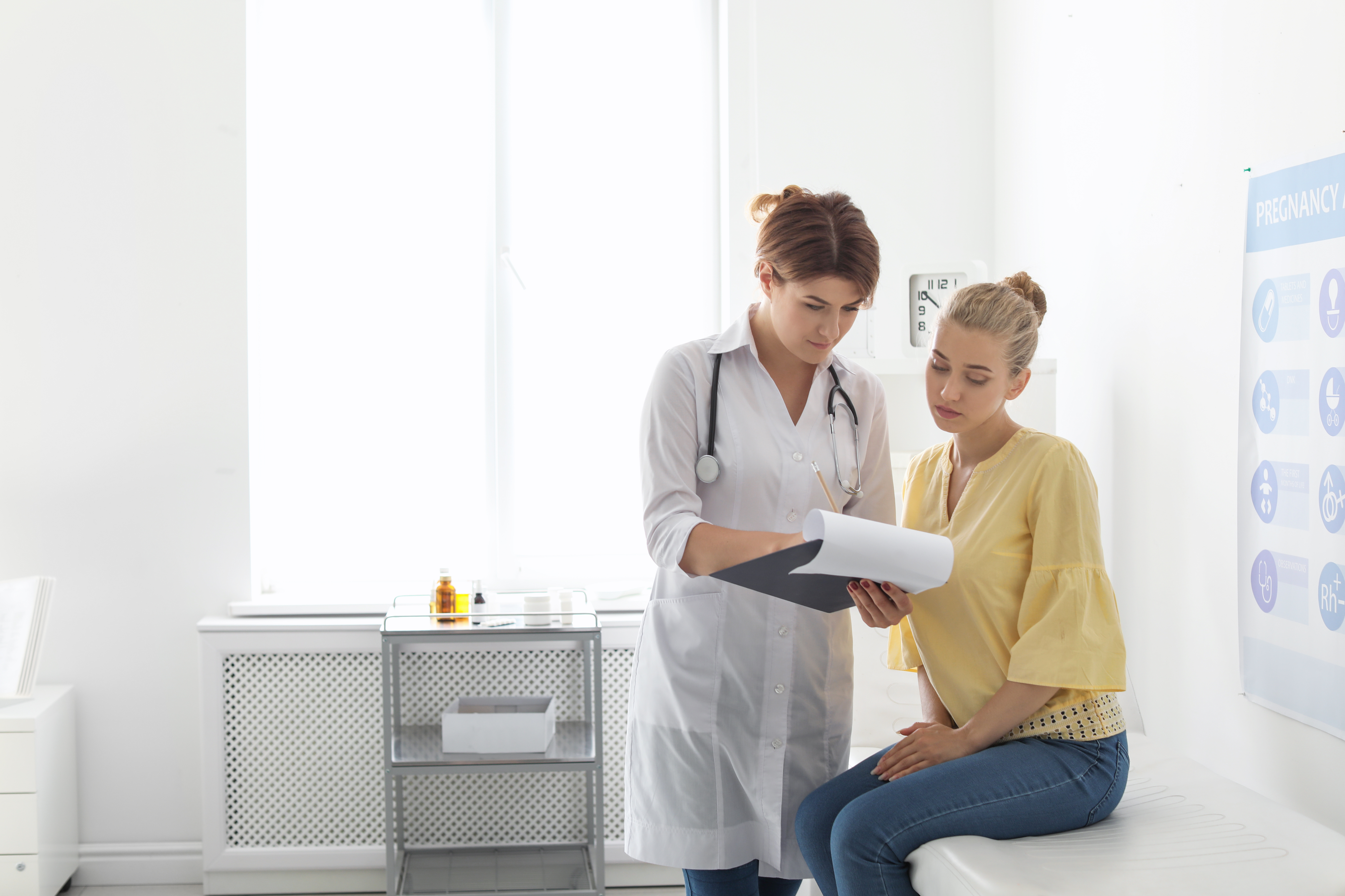 Young woman sitting on exam table at doctor's office with female doctor, looking at paperwork