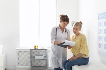 Young woman sitting on exam table at doctor's office with female doctor, looking at paperwork
