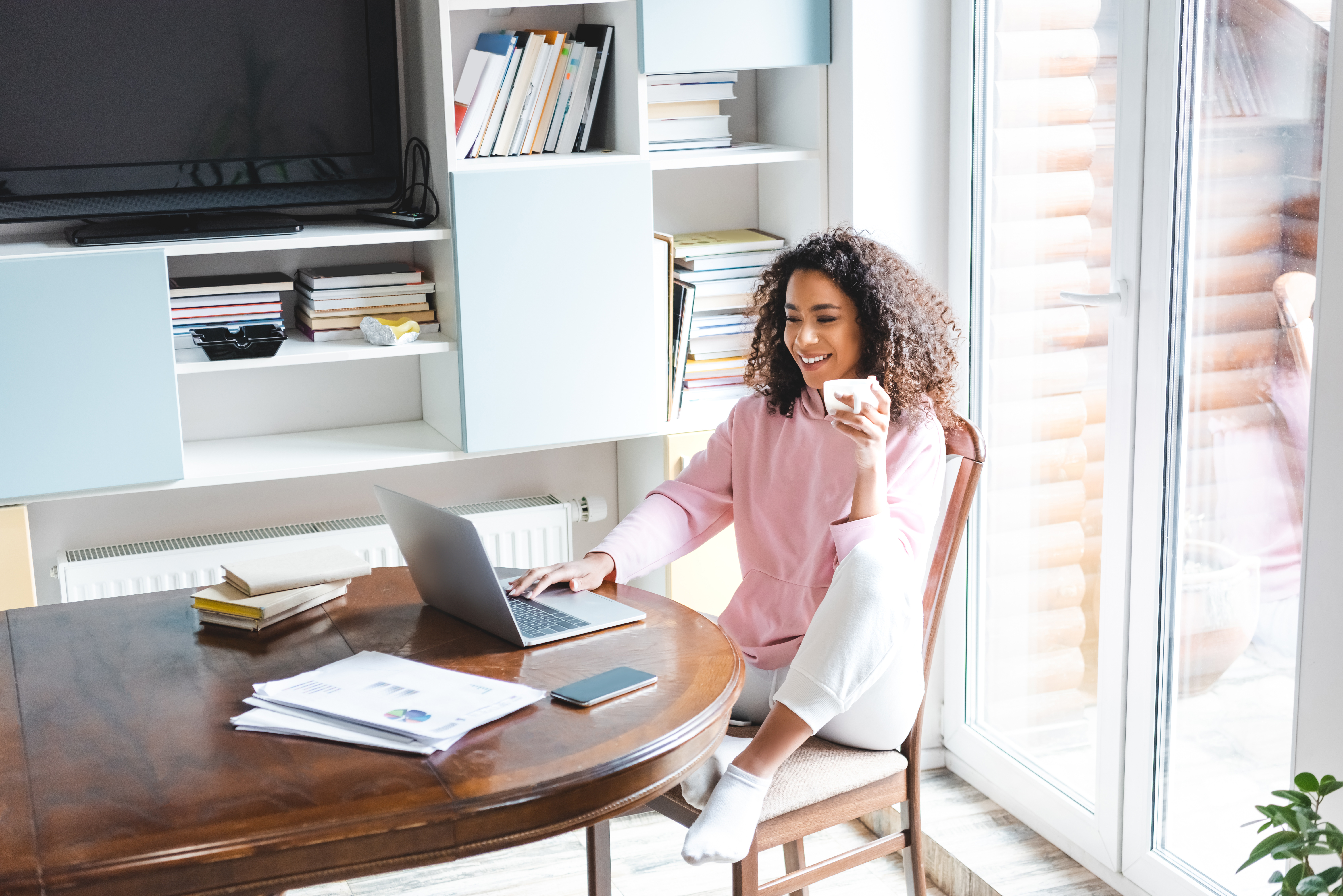 A young Black woman wearing a pink sweater and white sweatpants is sitting at her dining room table, drinking coffee while looking at her laptop.