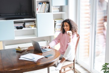 A young Black woman wearing a pink sweater and white sweatpants is sitting at her dining room table, drinking coffee while looking at her laptop.