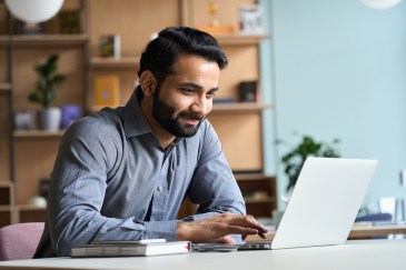 A man in a blue button-up is smiling at his silver laptop in his living room. The wall is blue and there is a wooden shelf behind him.