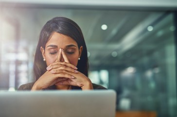 A South Asian woman is in front of her laptop, her eyes closed and fingers pressed to her forehead in exhaustion.