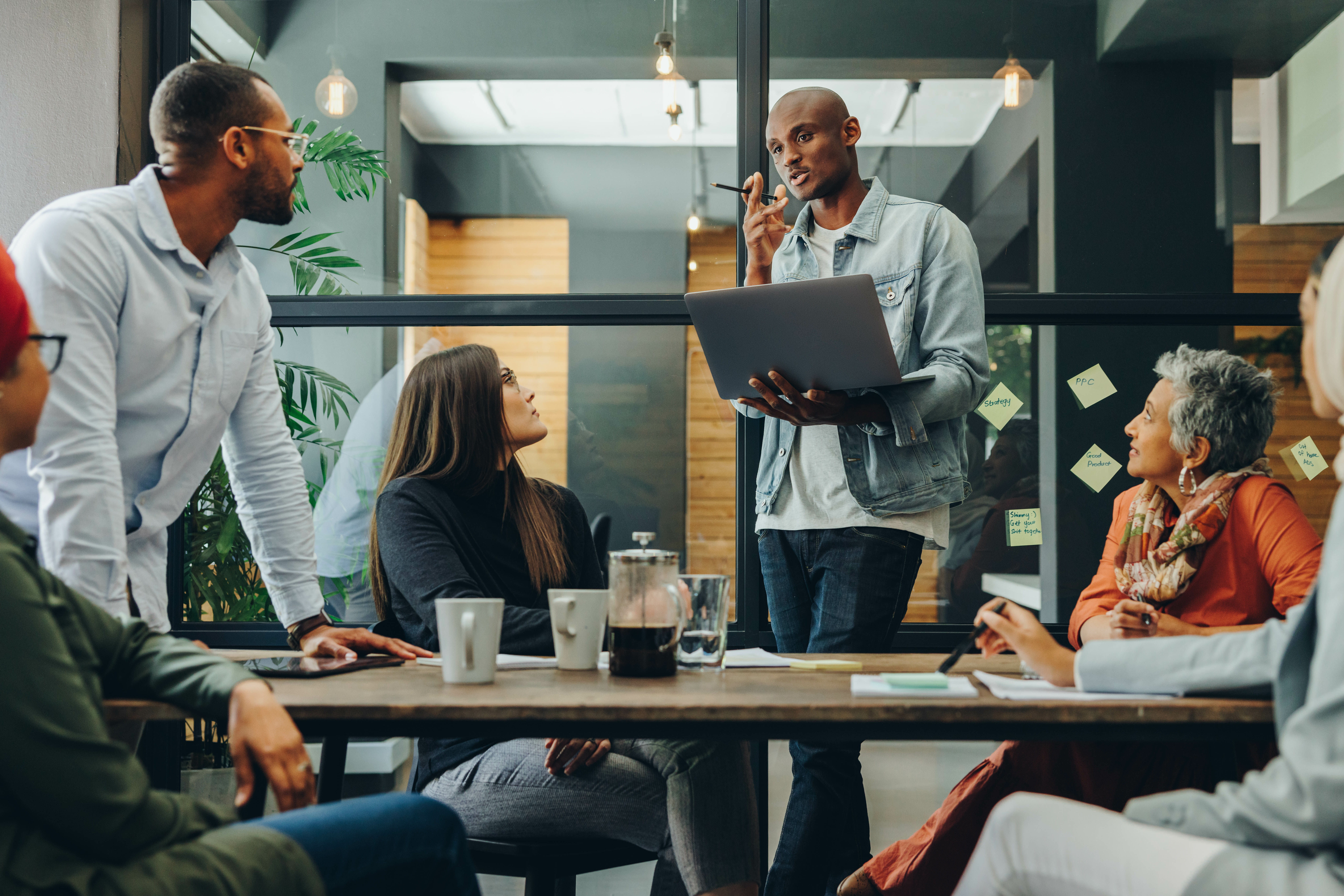 Six people sit around a table at their office; one Black man is explaining something to the group with a laptop in his hand.