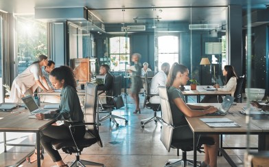 A modern office, with wooden desks in an open space and workers either sitting or walking through the space.