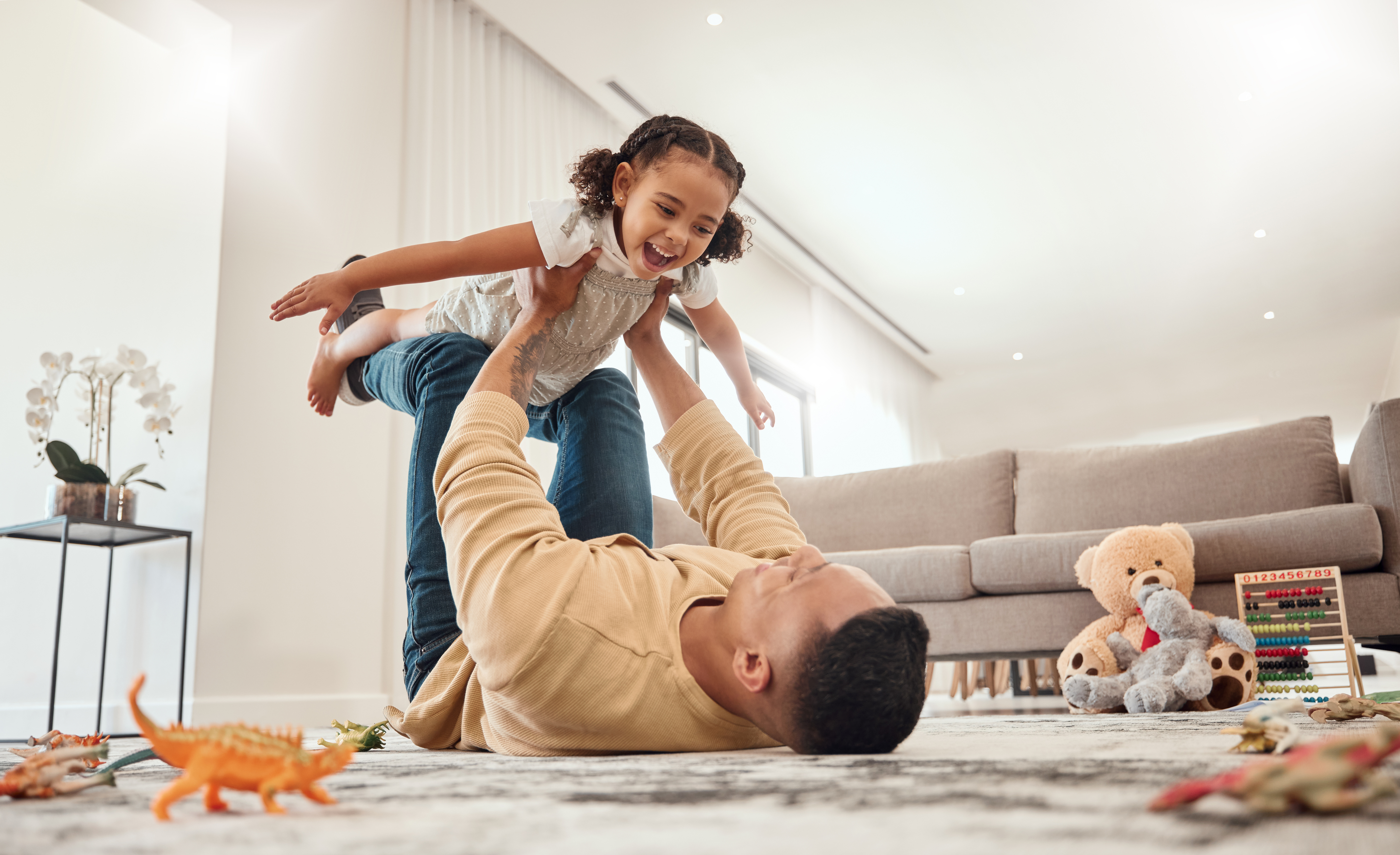 A dad is lying on the floor while holding his toddler daughter in the air; they are surrounded by toys like dinosaurs and stuffed animals.