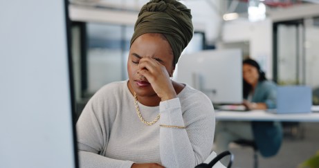 A Black woman in front of a desktop computer has her hand between her eyebrows and eyes closed in frustration.