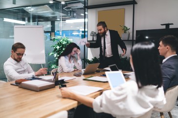 An angry boss yells at four employees who are seated around a wooden table inside a glass conference room.