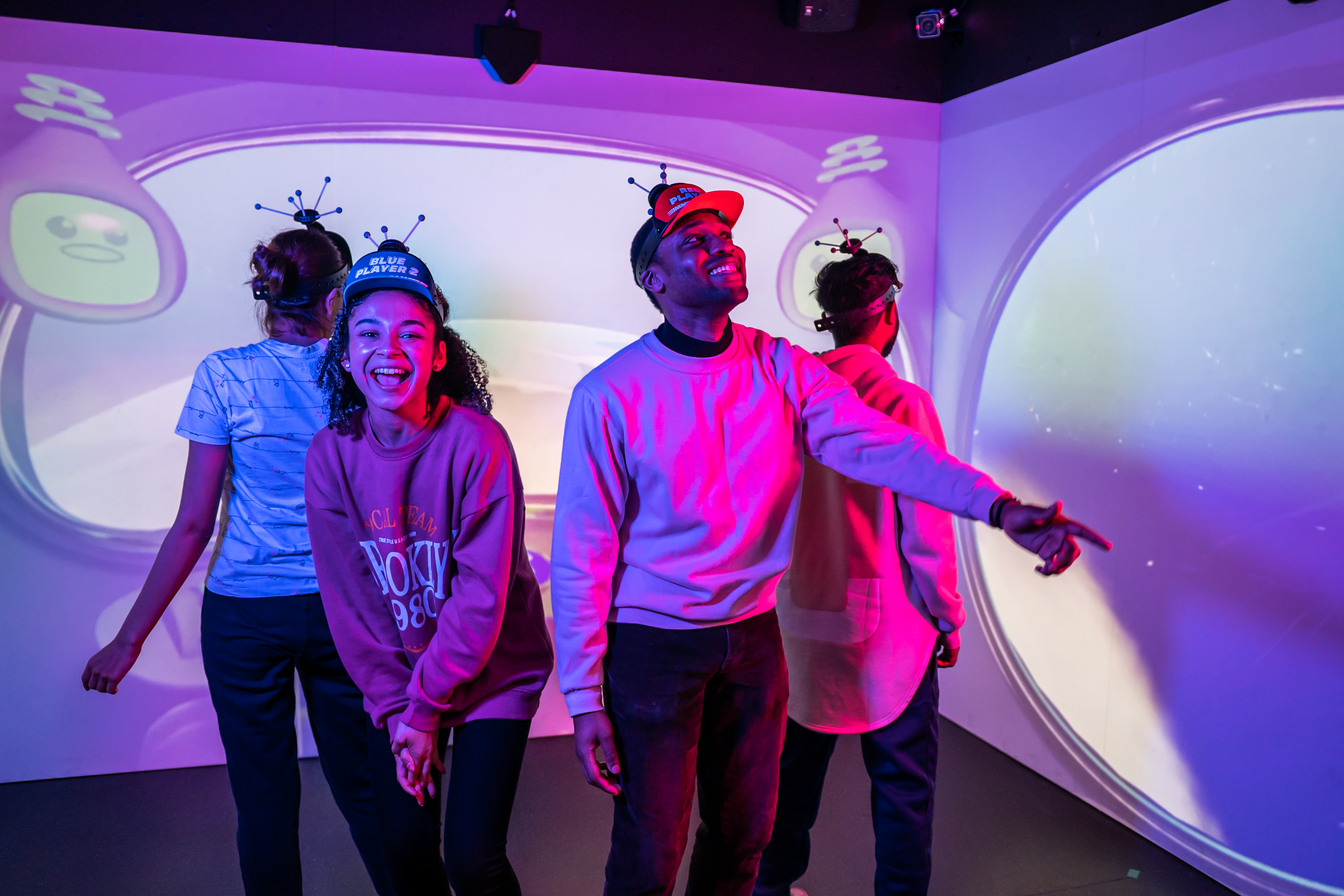 A group of four friends with visors on their head are looking around a digital game room -- a room filled with screens with cartoon characters on it.