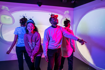 A group of four friends with visors on their head are looking around a digital game room -- a room filled with screens with cartoon characters on it.