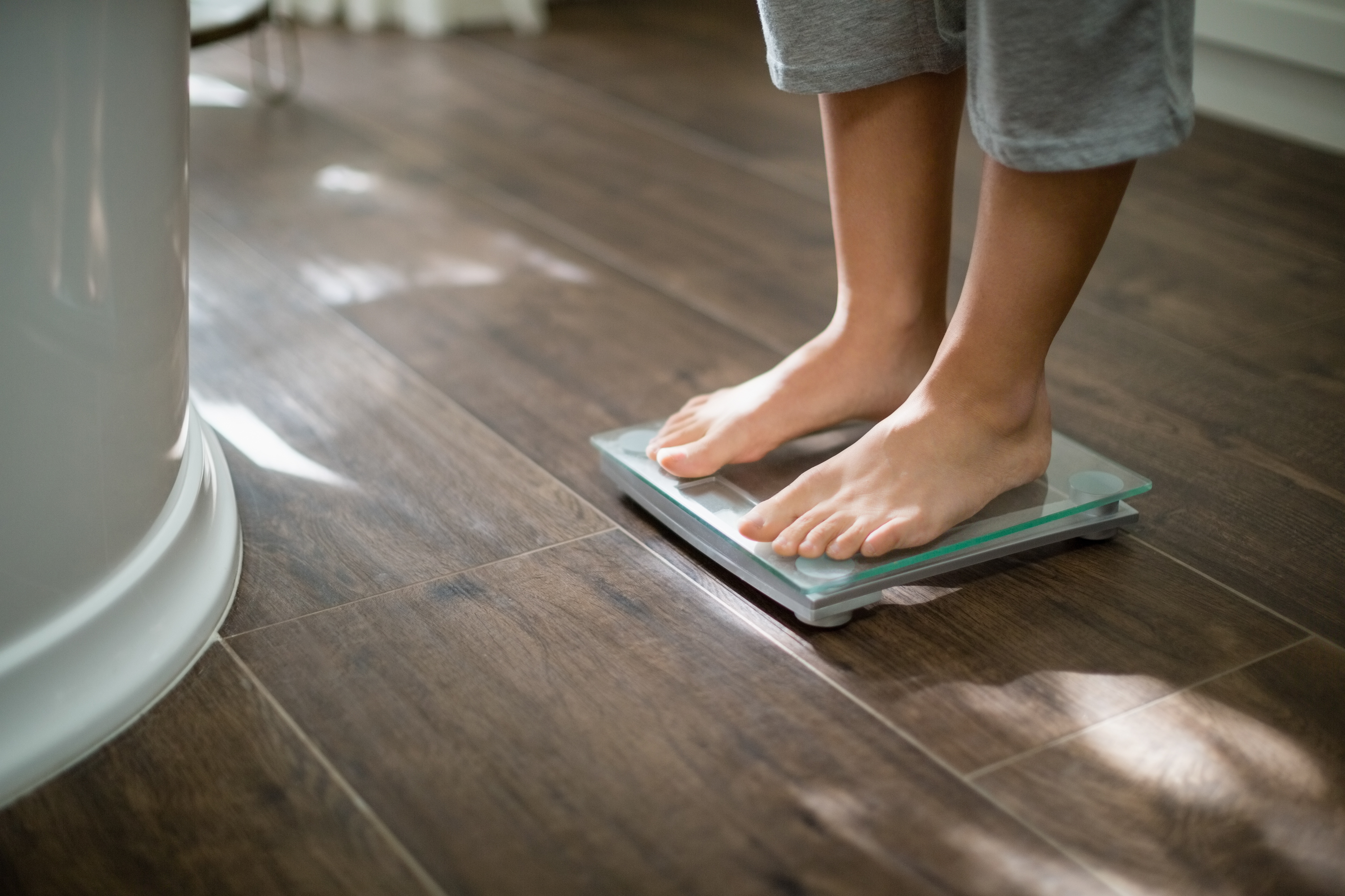 A person, barefoot, stands on a scale in their bathroom.