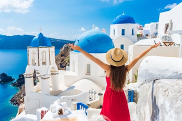 A woman in a sun hat and red dress has her arms spread out before a blue-domed church on the Italian coast.