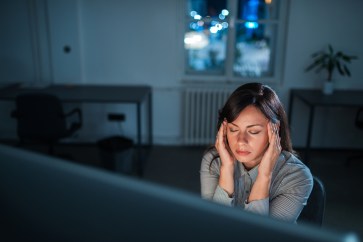 A woman rubs her temples in front of her desktop computer; the room is dark, with blue light cast off the computer.