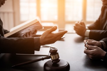 Men sitting on opposite sides of table; one holding book; judge's gavel in between