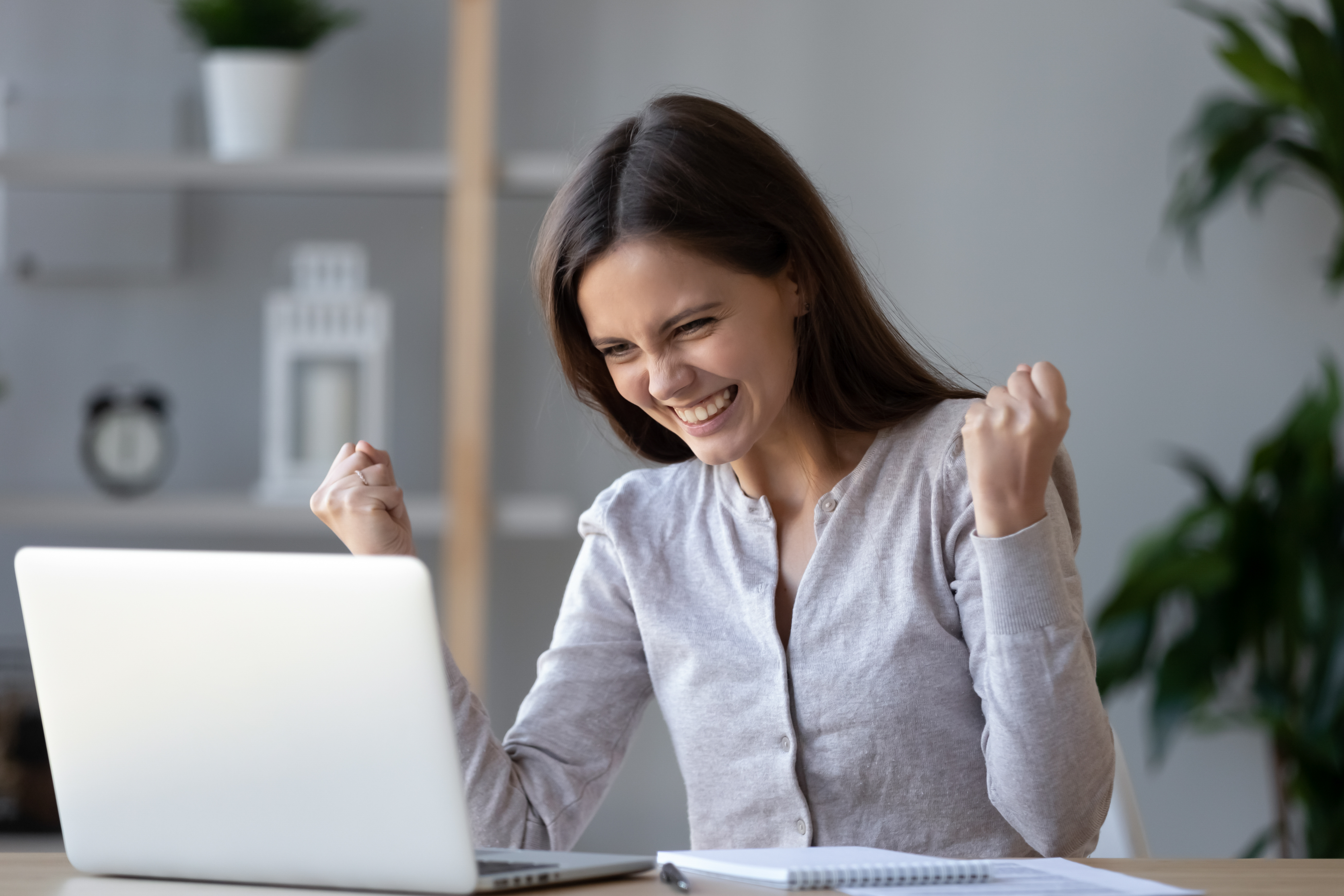 A brunette, white woman puts both her fists in the air in celebration while looking at her laptop.