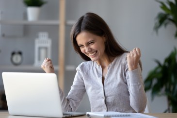 A brunette, white woman puts both her fists in the air in celebration while looking at her laptop.