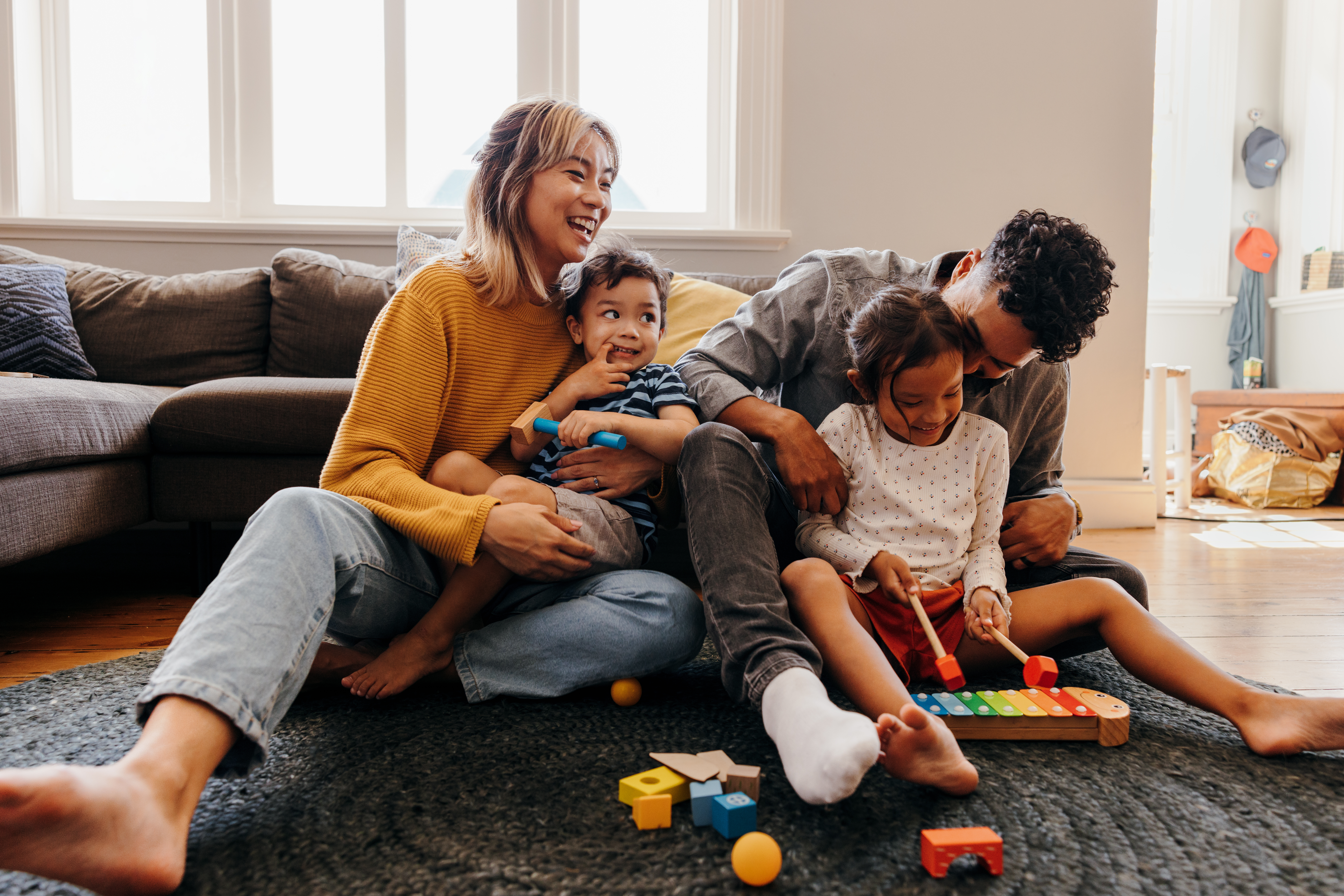 Parents with a son and daughter are holding them in their laps while laughing in their living room; there are colorful toys on the rug at their feet.