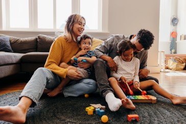Parents with a son and daughter are holding them in their laps while laughing in their living room; there are colorful toys on the rug at their feet.