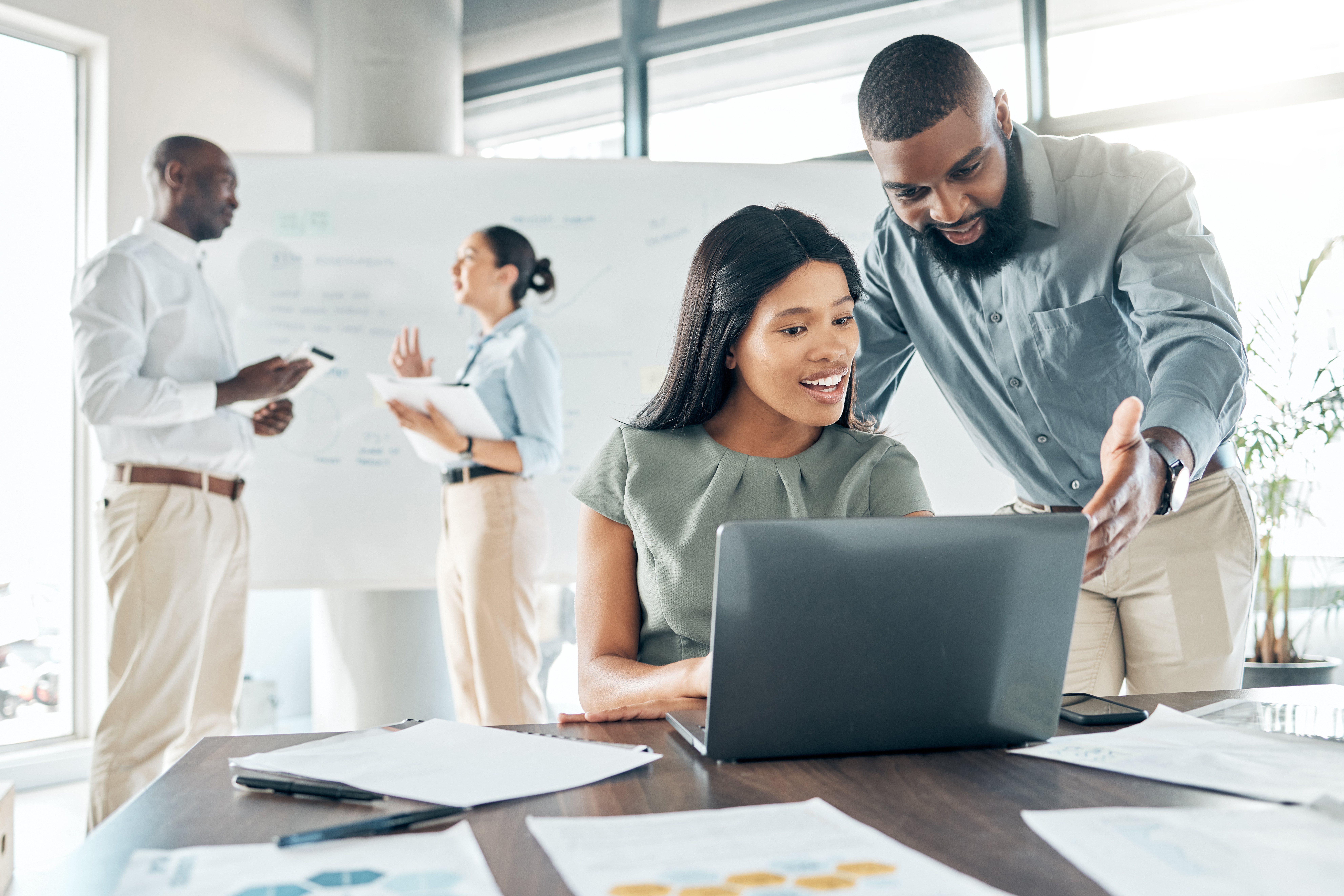 Employees working on laptop, one standing and motioning to computer screen; one sitting and smiling