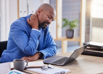 A Black man in a blue suit is rubbing his neck, with his eyes closed at his desk and his laptop open.