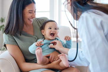 A mom is sitting with her baby while a doctor listens to the baby's heartbeat with a stethoscope.