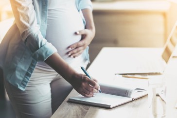 A pregnant woman touches her stomach while writing something down in her notebook at her desk.