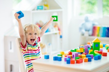 A little kid, around 5 years old, excitedly throws up their arms with colorful bocks in each hand. The table in front of them has a pile of building blocks.