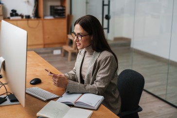 Woman with glasses in office in front of computer holding phone