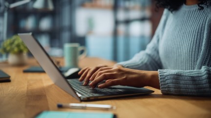 A woman in a blue knitted sweater is typing on her laptop on a wooden table.