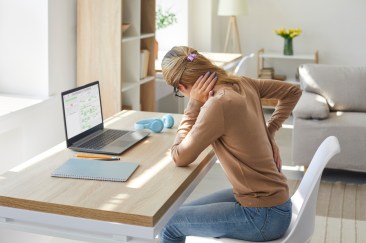 A woman rubs her neck and has her other hand on her lower back; she sits at her desk in her sunlit living room, turned away from the camera in discomfort.