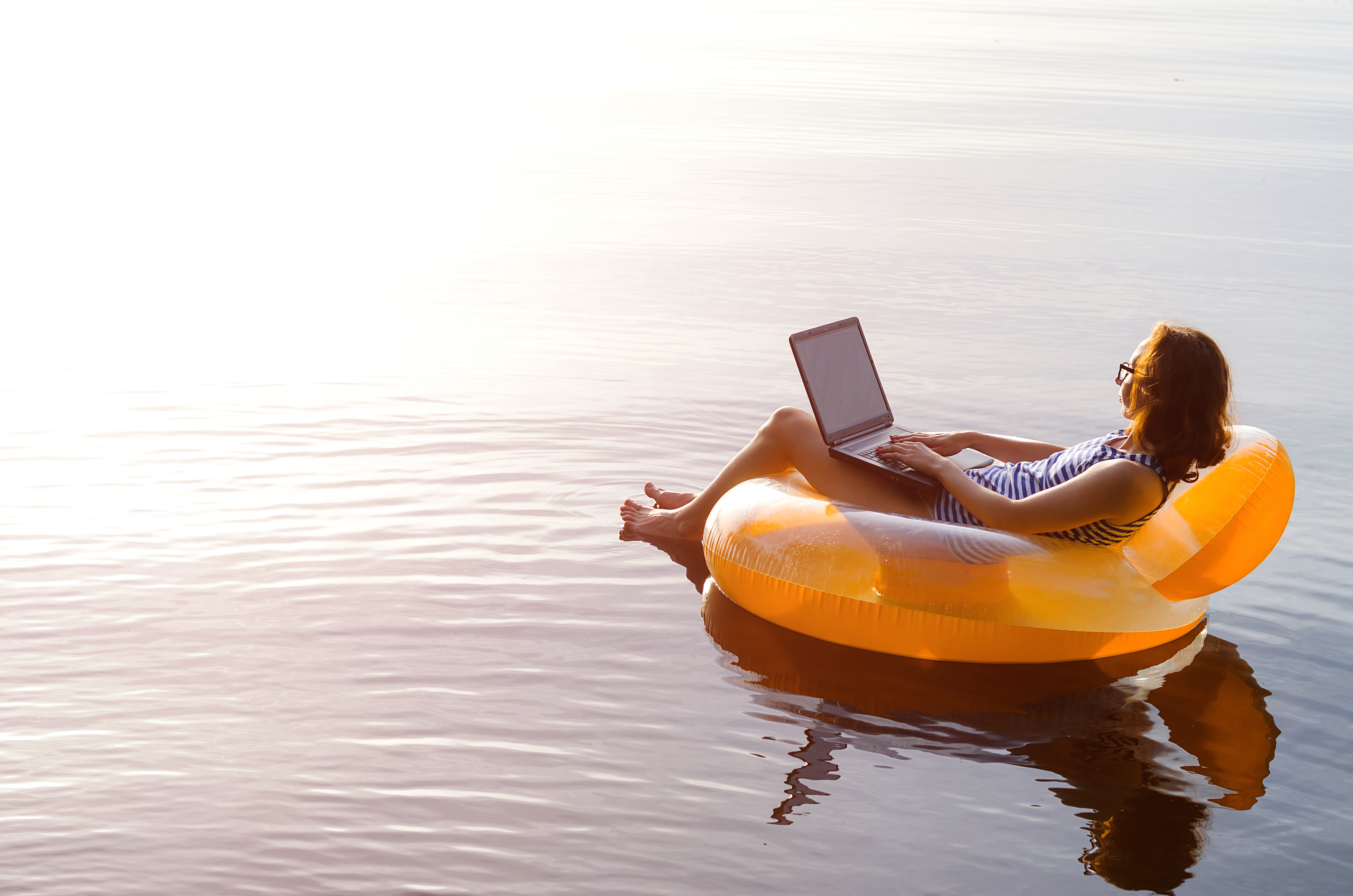 A woman is floating in an orange tube on the lake while on her laptop.
