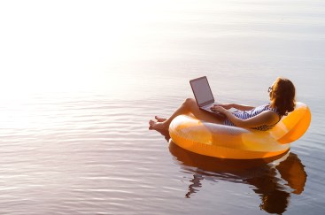 A woman is floating in an orange tube on the lake while on her laptop.