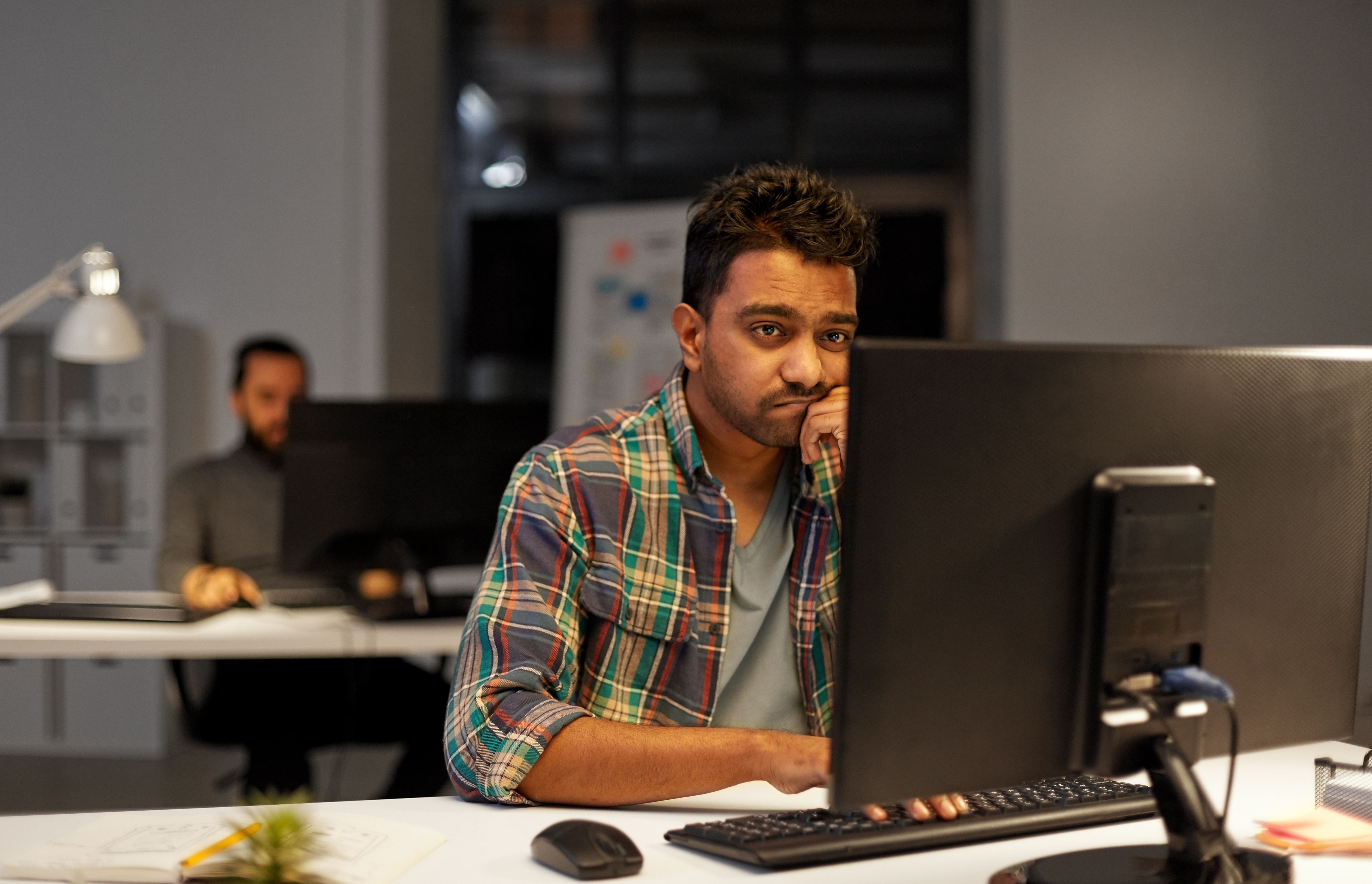 A young worker looks sadly at his desktop computer in an open-layout office; it's the evening.