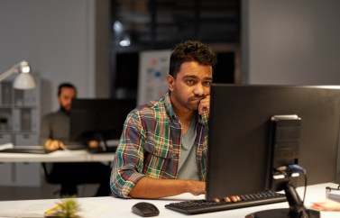 A young worker looks sadly at his desktop computer in an open-layout office; it's the evening.