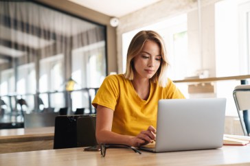A woman is working on her laptop in a sunlit room with other desks behind her.