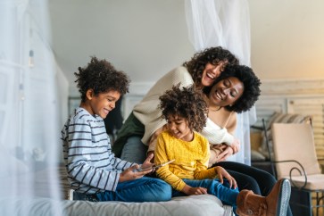 Two moms smile as their two kids (son and daughter) laugh at something on an iPad.
