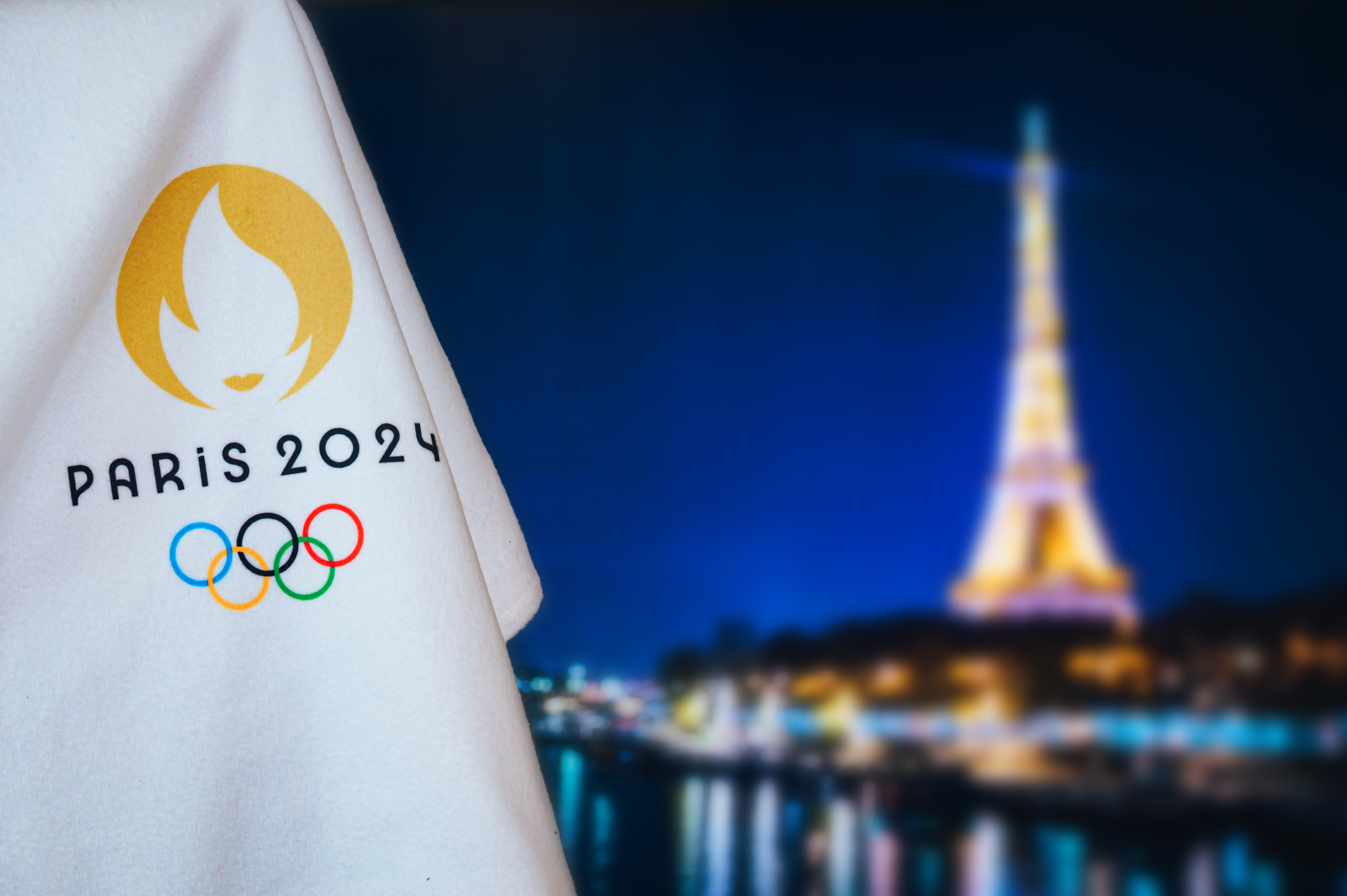 A flag that says "Paris 2024" with the Olympic rings is on the left side of the frame, with the Eiffel Tower in the background.