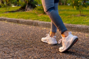 A woman with white tennis shoes, which take up the frame, is walking along a path alongside grass.