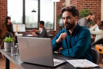 Man sitting in office looking at laptop with pen up to mouth, thinking