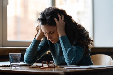 A woman has her hands in her hair in frustration as she looks down at her desk.