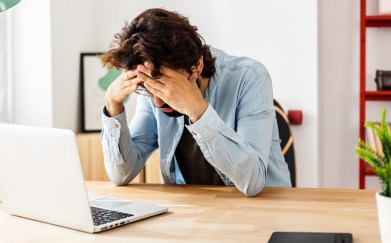 A man puts his head in hands while looking down at his laptop.