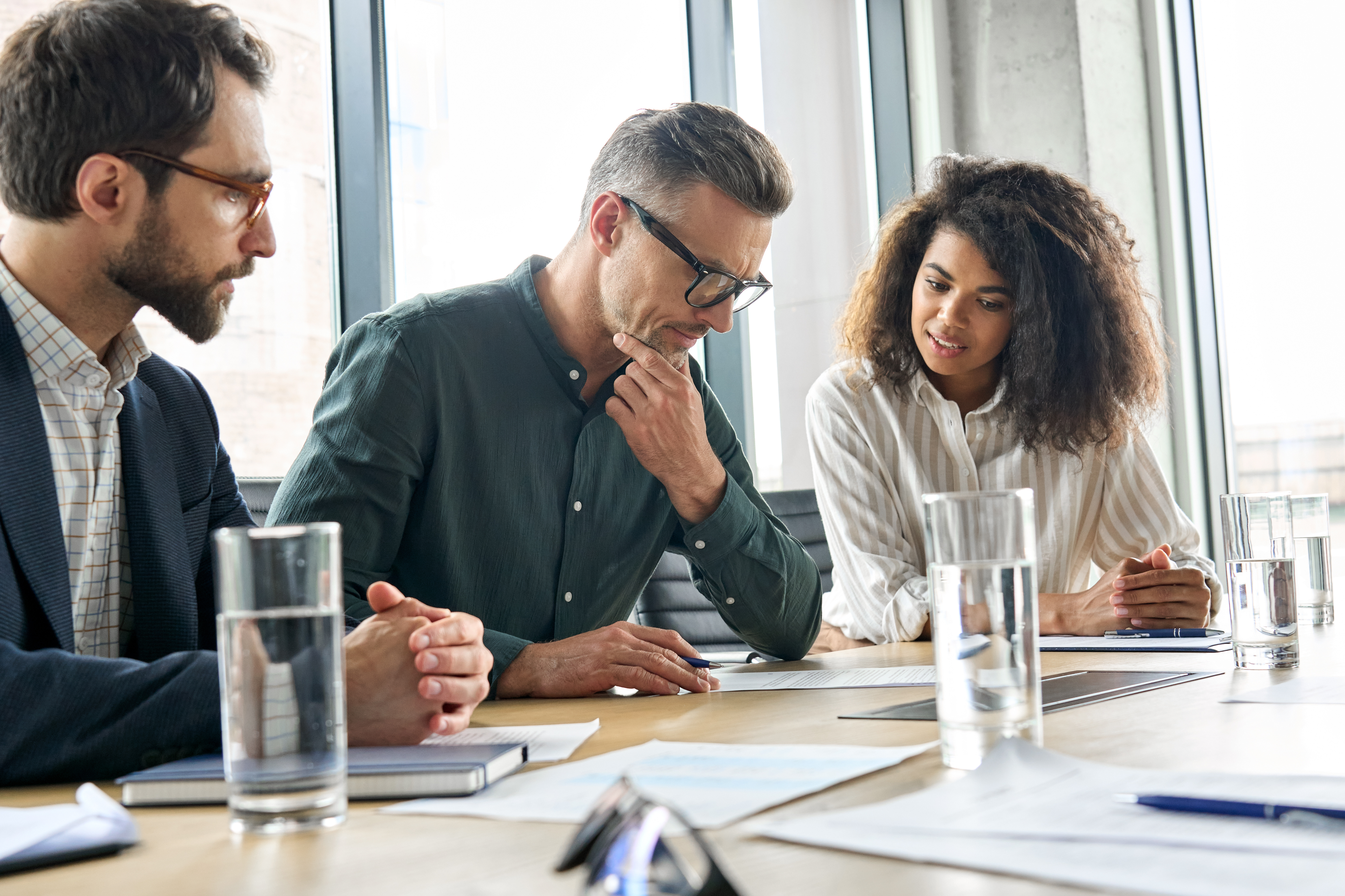 Three corporate employees sitting at a conference stare down at a piece of paper, looking concerned.