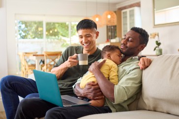 Two dads hold their baby daughter while sitting on a couch in a sunlit living room.