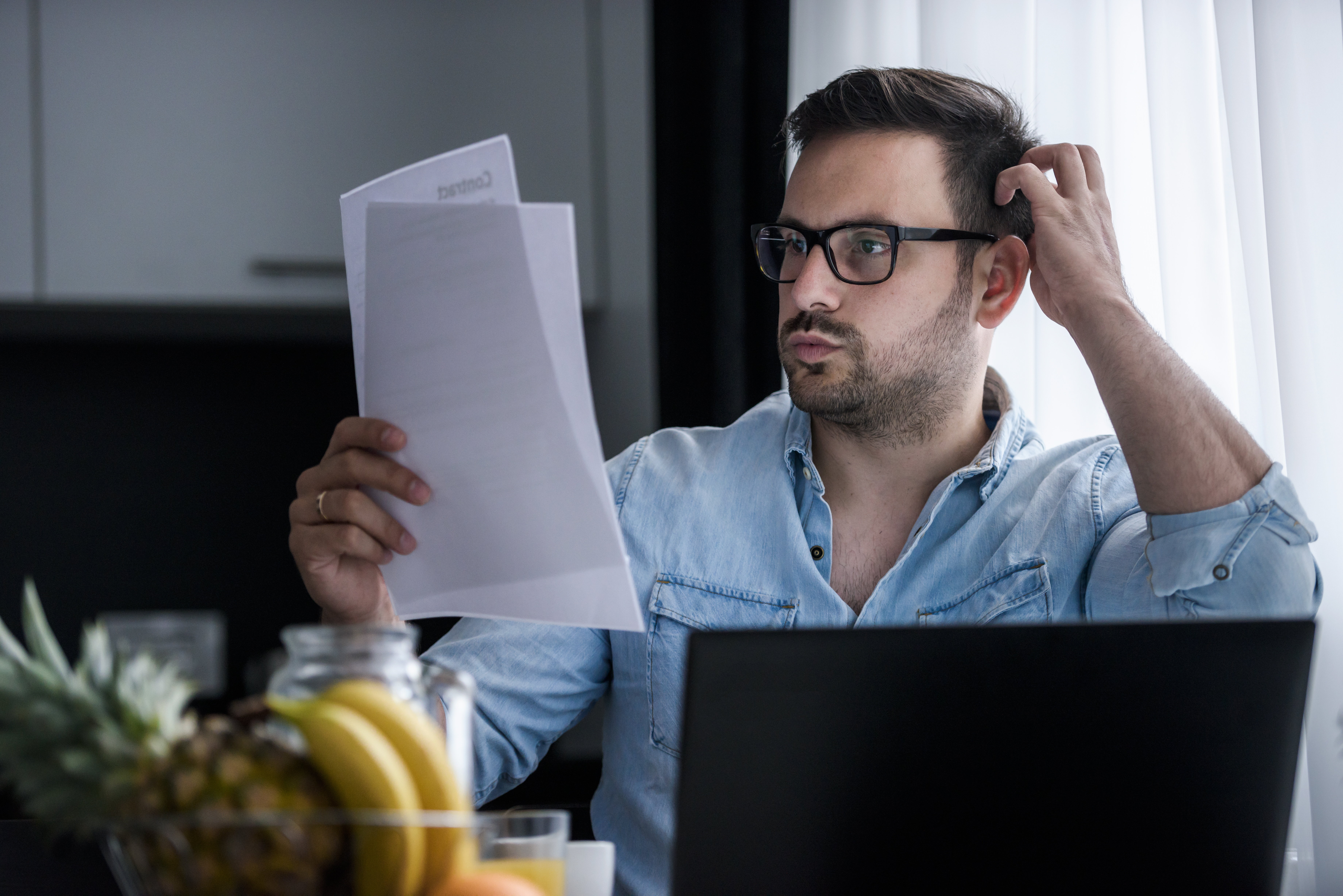 Man looking at paperwork sitting at table with computer, looking confused