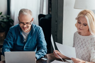 Man and woman sitting at table with laptop and papers; reading paperwork