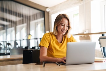 A woman is smiling at her laptop while the sun shines through the window behind her.