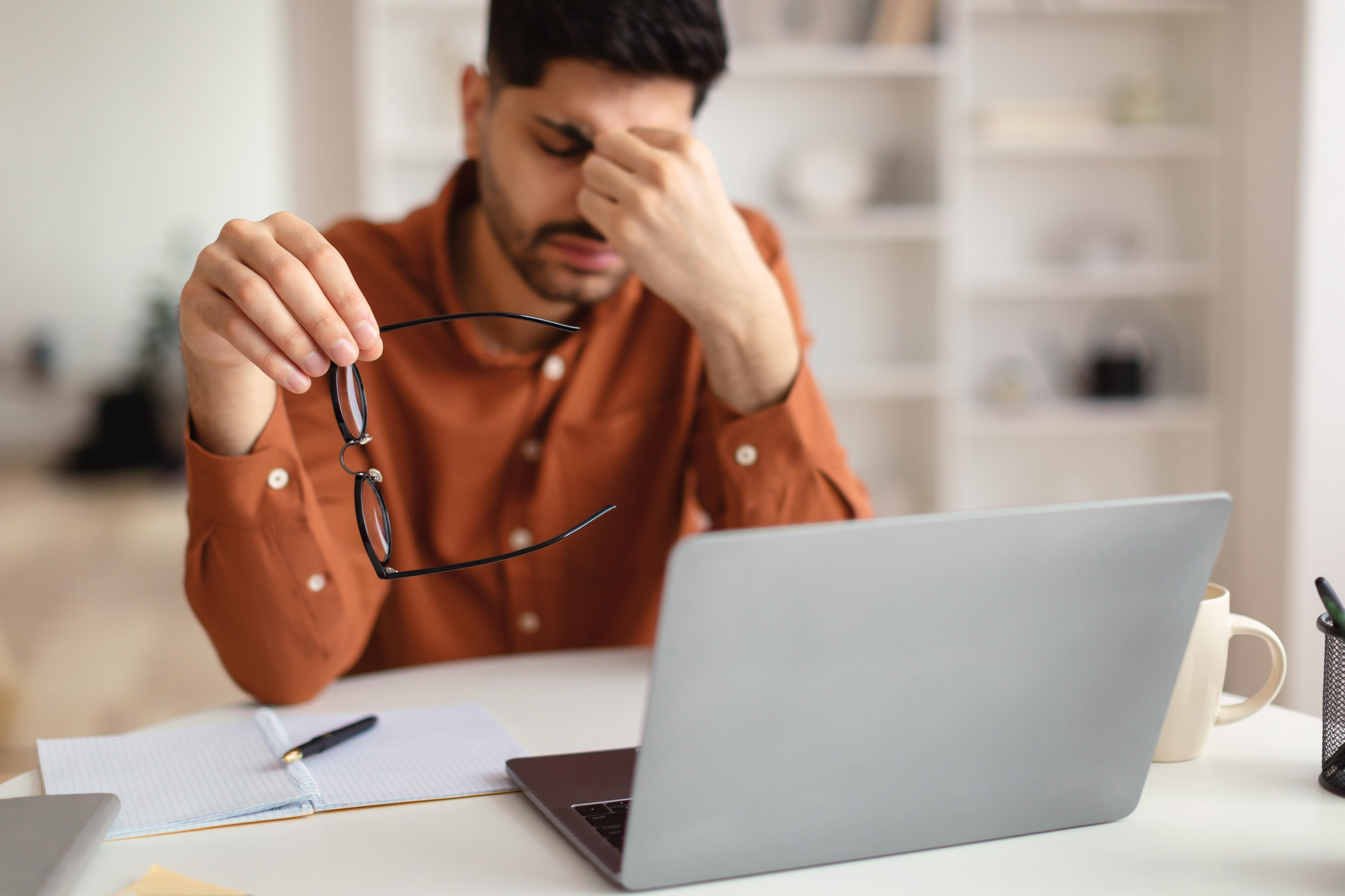 Man sitting in front of laptop, holding glasses in hand and pinching nose, stressed