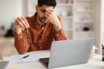 Man sitting in front of laptop, holding glasses in hand and pinching nose, stressed